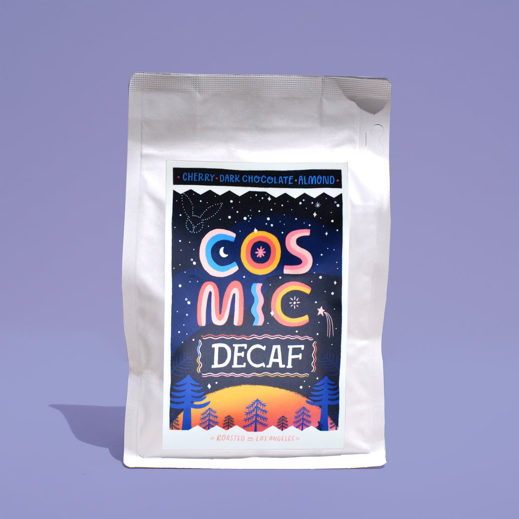 A side view of a white bag of coffee on a purple backround. The label is blue with a starry sky illustration. The copy reads "COSMIC DECAF""