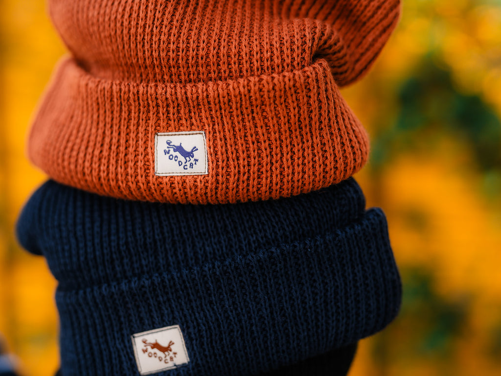 A close-up shot of two knit beanies stacked on top of one another. The top beanie is an organe-brown color and the bottom beanie is a navy blue. Both beanies have small rectangular white tags sewn to the side that read "woodcat"