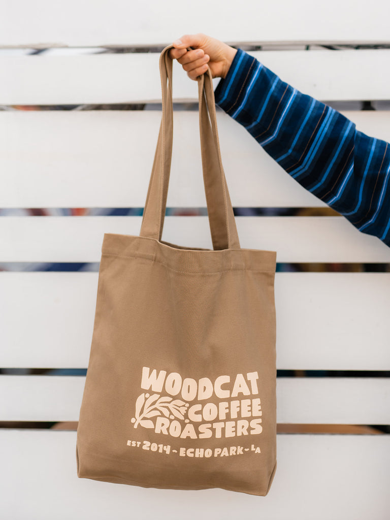 An off-screen arm holding a large tan tote bag. It reads "woodcat coffee roasters" in large cream block letters.