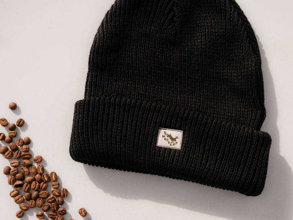 Black cable knit beanie on a white background with a small array of coffee beans off to the left side. The beanie has a small white square tag with an embroidery in brown that reads "woodcat"