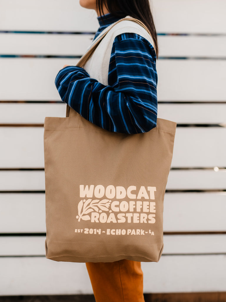 A female-figure standing in front of a white horizontal fence. She is facing to the left and has a large tan bag over her shoulder as left arm is holding the strap. She is wearing a black and blue striped long-sleeved shirt with a white knit sweater vest. The tote bag reads "woodcat coffee roasters" in large beige block letters.