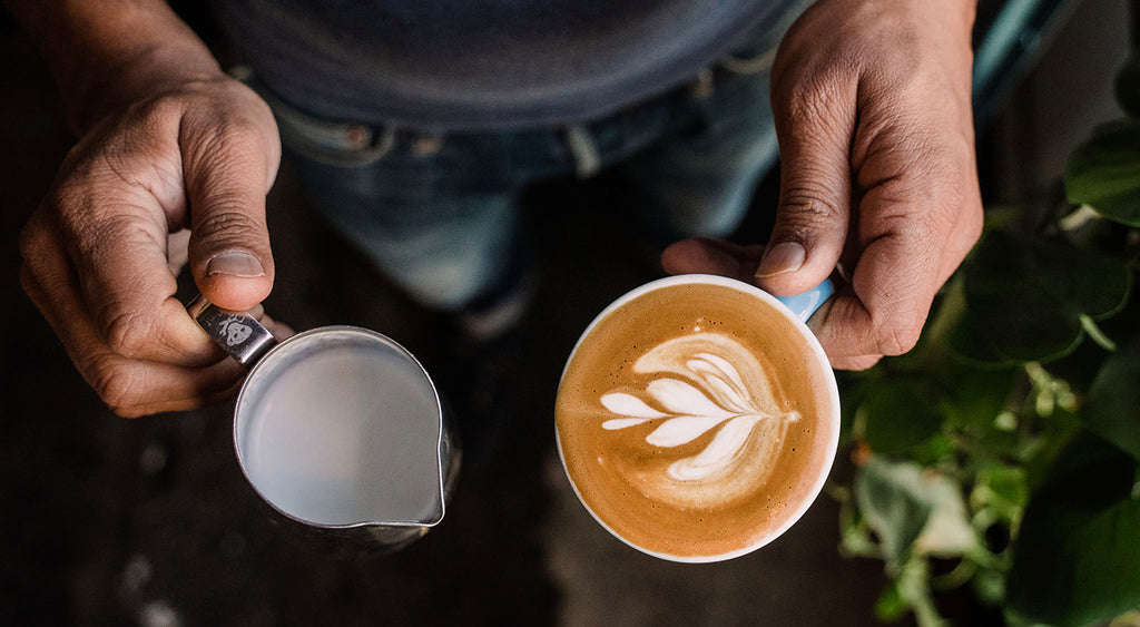 An overhead shot of two hands. The left is holding a milk pitcher and the right hand is holding a cappucino cup with white latte art