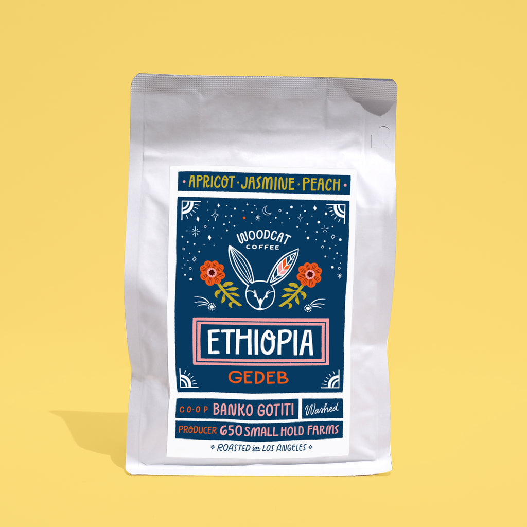 A white coffee bag with a dark blue label on a yellow backdrop. The label reads "Apricot, Jasmine, Peach. Woodcat Coffee. Ethiopia Gedeb"