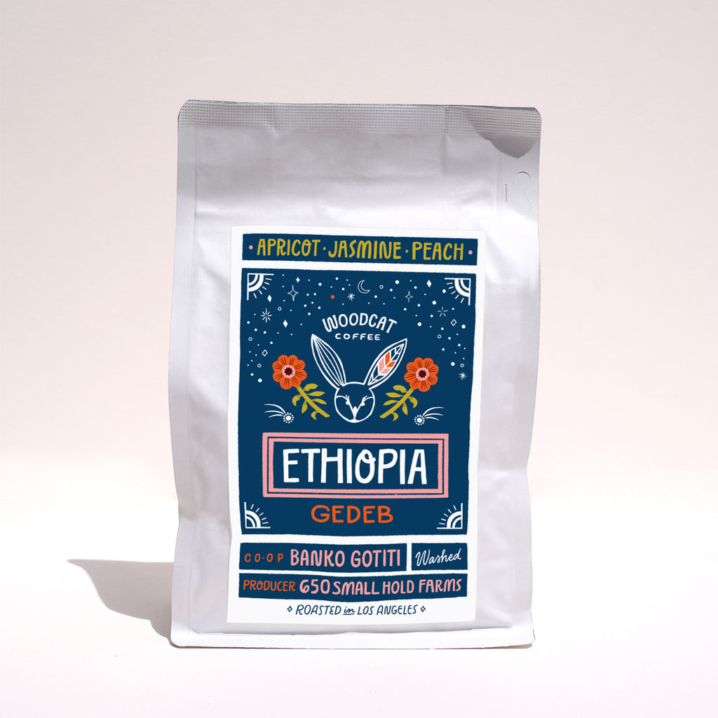 A white coffee bag with a dark blue label on a white backdrop. The label reads "Apricot, Jasmine, Peach. Woodcat Coffee. Ethiopia Gedeb"