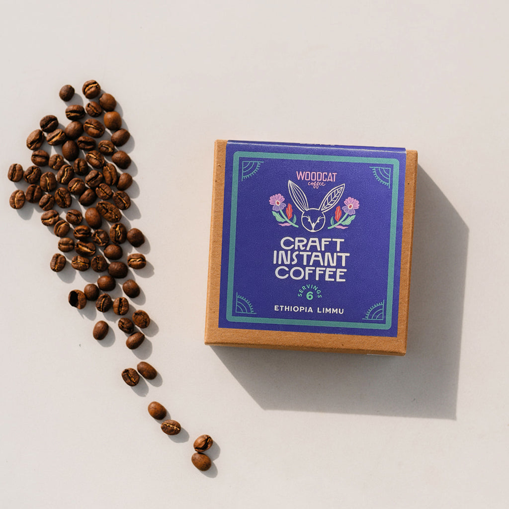 A photo of a brown paper color box with a blue label that reads " Woodcat Coffee. Craft Instant Coffee. 6 Servings. Ethiopia Limmu." Coffee beans are scattered to one side of the box