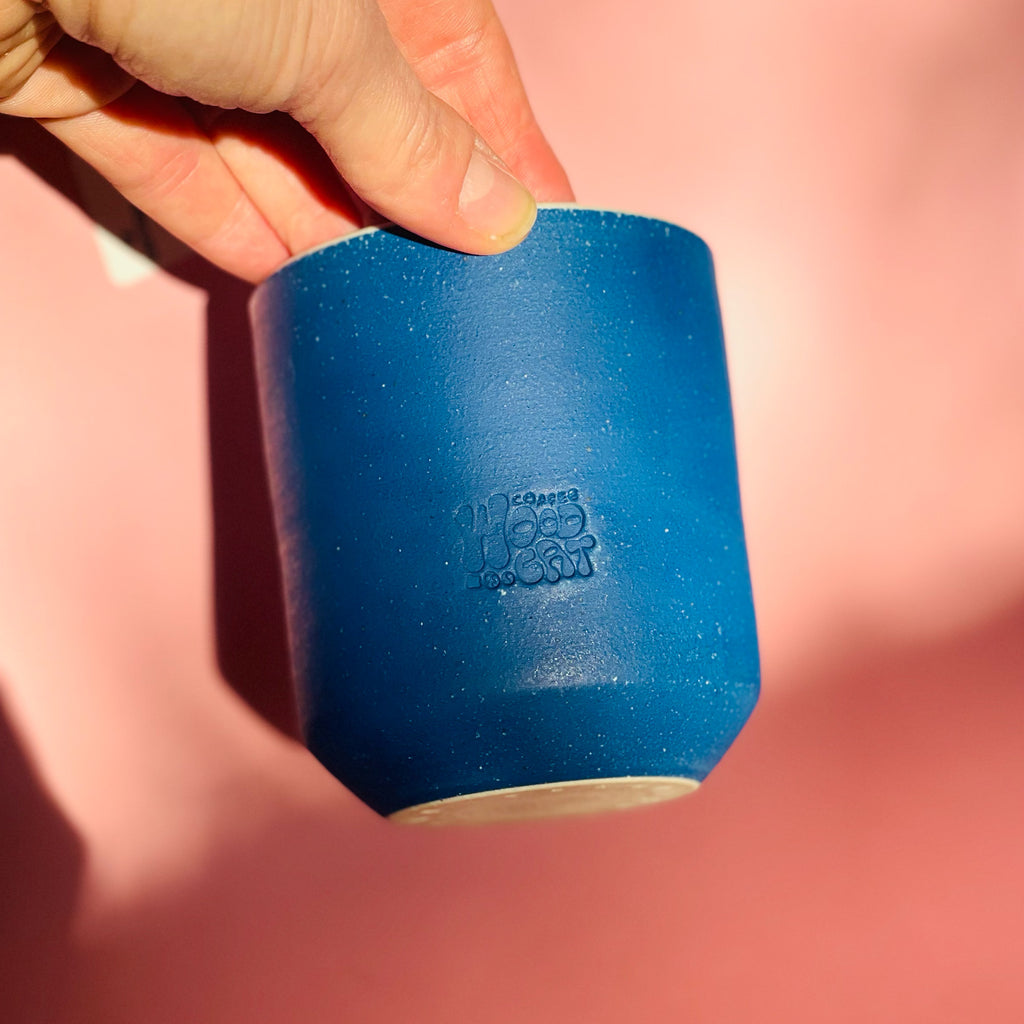 a smooth blue cylindrial vessel with a embossed "woodcat" logo. Someone is holding the vessel from the top infront of a pink background.