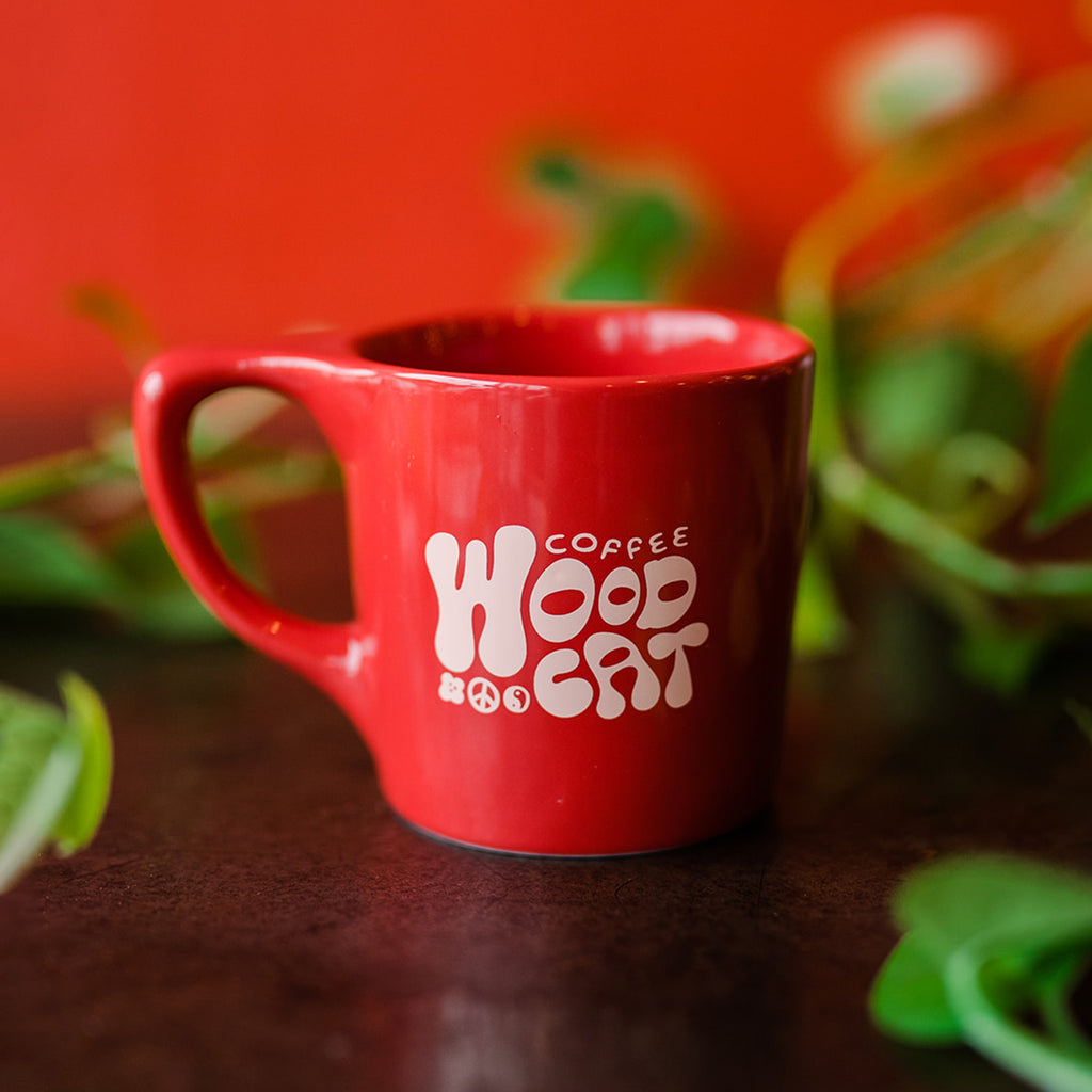 a red mug with pink bubble lettered design. It is sitting atop a dark colored table and is surrounded by green foliage..